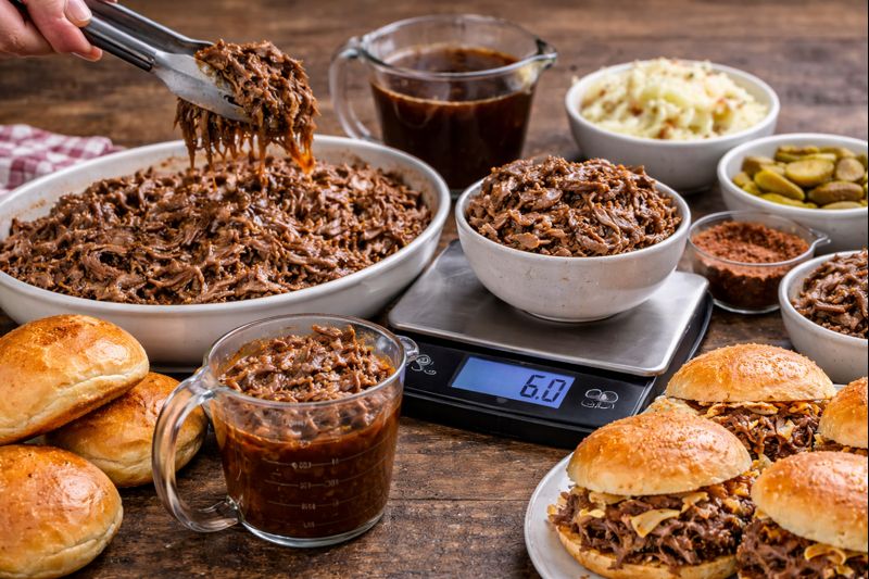 Shredded beef being portioned with tongs beside sandwich buns and sides to show serving sizes per person